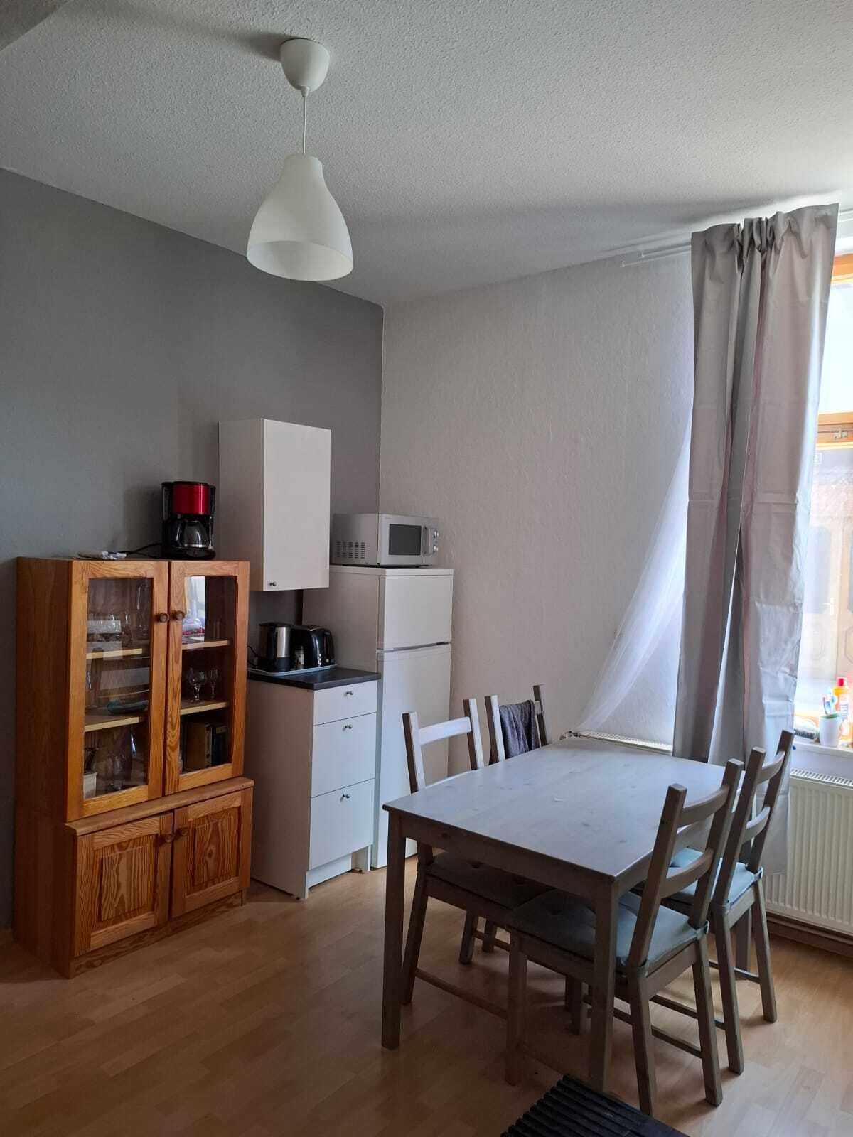 Eine kompakte Küche mit Essbereich, Holzschränken, einer weißen Deckenleuchte und einem Fenster mit Vorhängen.