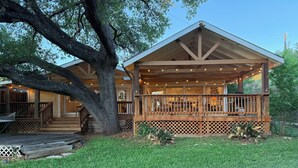 A wooden cabin with a covered porch, a staircase leading to the entrance, and a large tree in the foreground. - Dog-Friendly Lakehouse: Dock + Serene Views (Burnet)