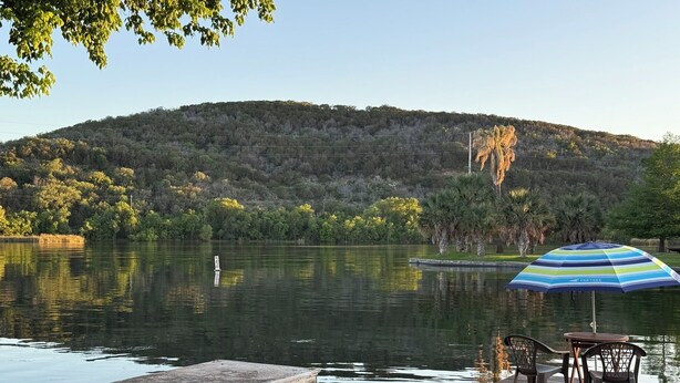 A lakeside dock with a table and chairs, an umbrella, and a view of a forested hill. - Dog-Friendly Lakehouse: Dock + Serene Views (Burnet)