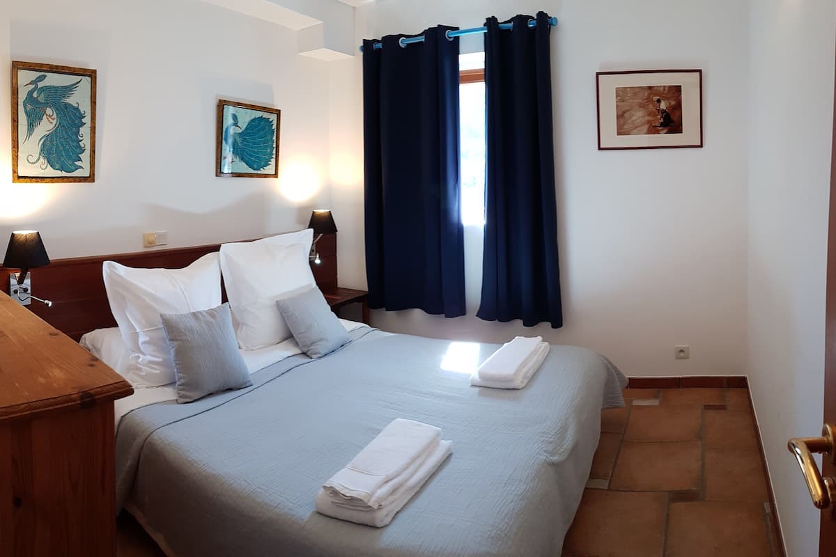Una cama bien hecha con almohadas y una toalla doblada, una mesita de noche de madera con una lámpara, y dos obras de arte enmarcadas en la pared.