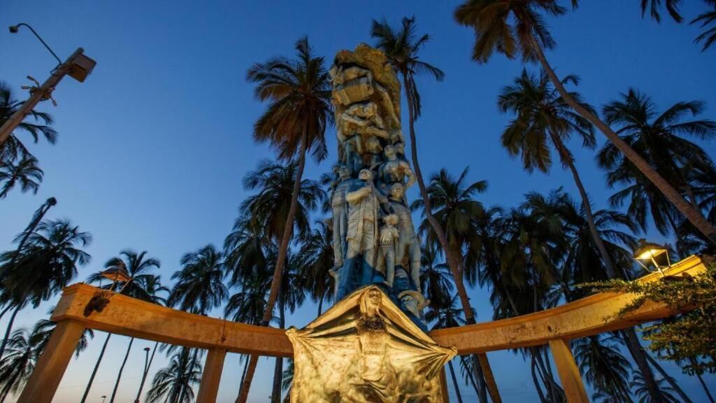 A tall, ornate statue with a figure at the top, surrounded by palm trees and a clear sky.