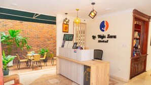 A hotel reception area with a wooden reception desk, a brick wall, and a reception desk with a logo. There are potted plants and a seating area with chairs and a table. - Hotel Galeria la Vieja Sara Riohacha (Riohacha)