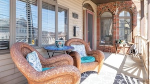 A covered porch with wicker chairs and a small table, a brick wall, and a window with a number sign.