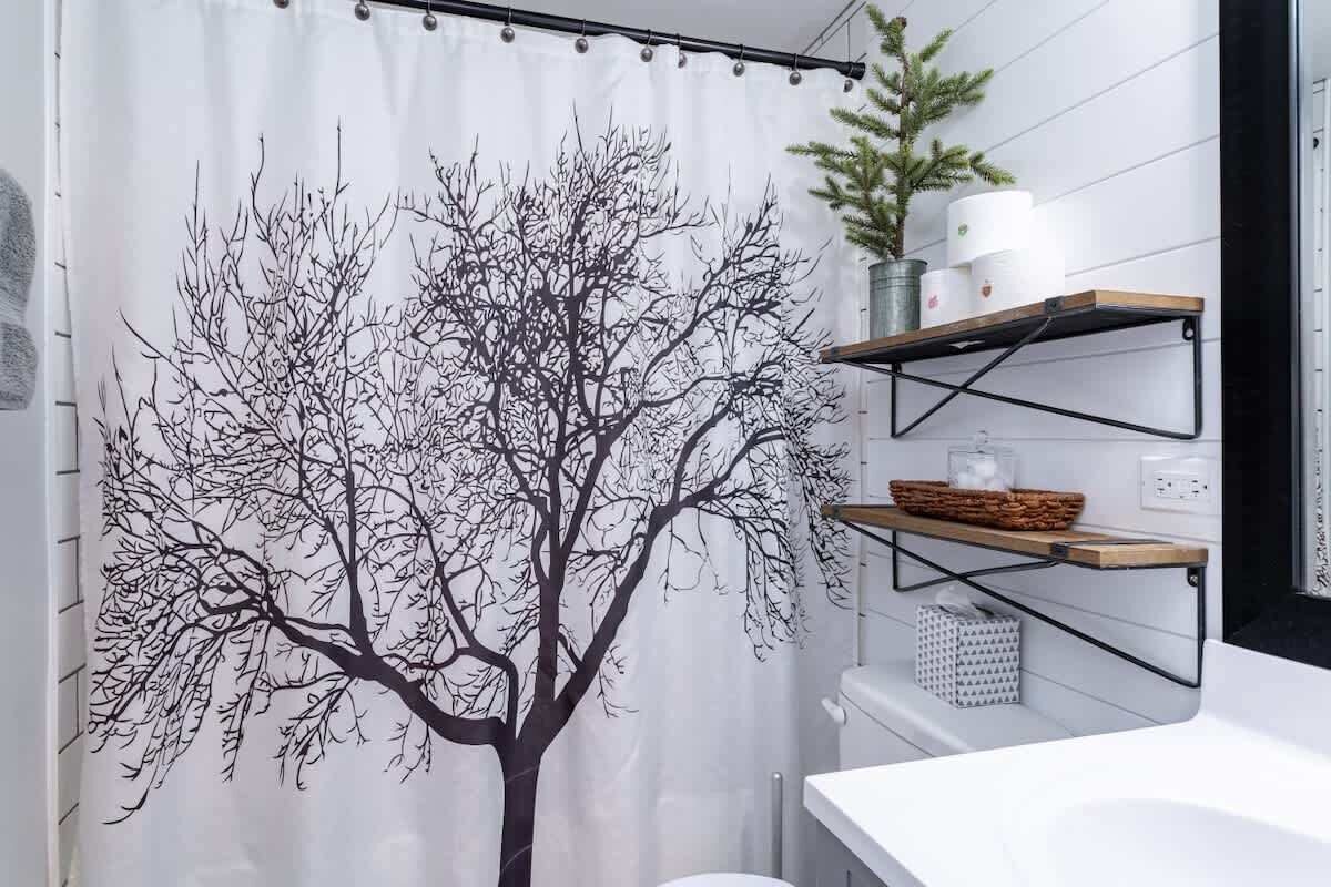 A bathroom with a tree decal on the shower curtain, a white sink, and shelves with toiletries.