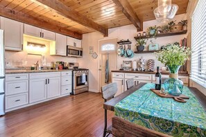 Une cuisine avec des meubles blancs, un plafond en bois, une table à manger recouverte d’une nappe à motifs et divers appareils de cuisine.
