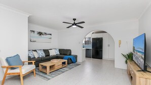 Un salon moderne avec un canapé, une table basse et un téléviseur. On y trouve également un ventilateur de plafond, un coin cuisine avec un réfrigérateur, et une porte qui donne sur une autre pièce.