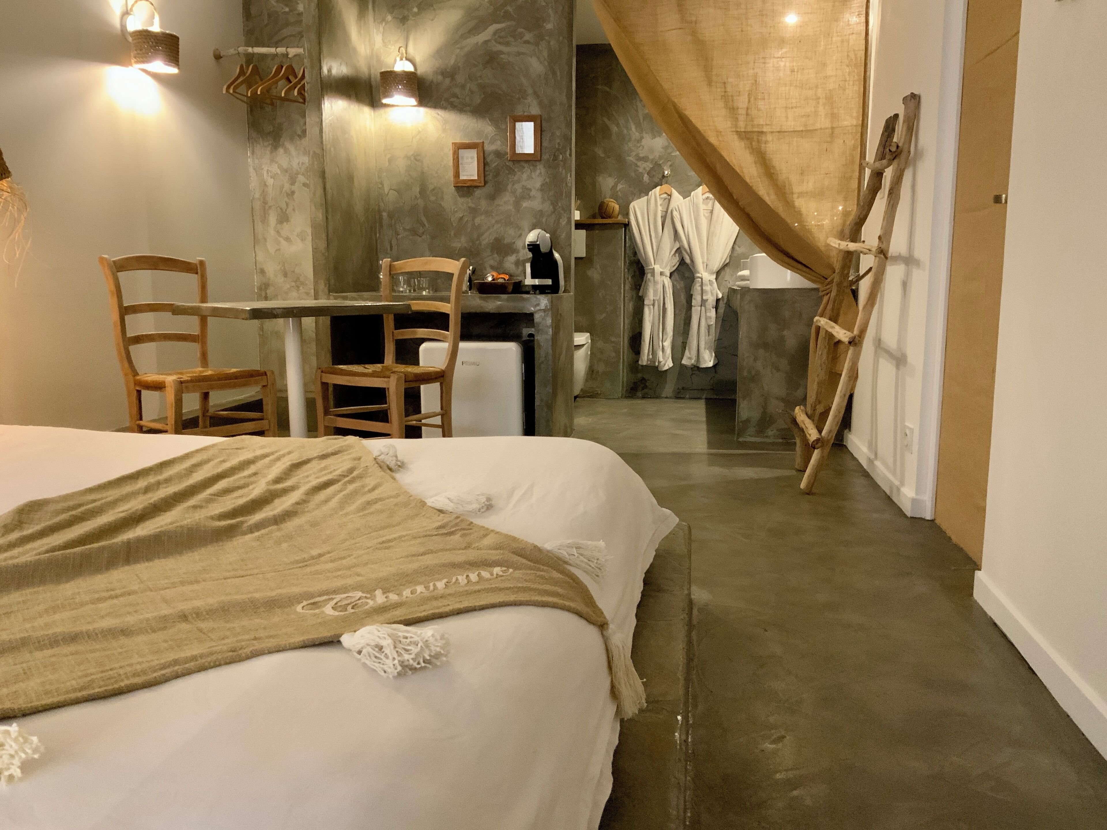 A hotel room with a bed, a desk, chairs, a small kitchenette, and a bathroom visible through a curtain.