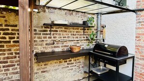 Een buitenkeuken met een bakstenen muur, een barbecue, planken en een hangplant.