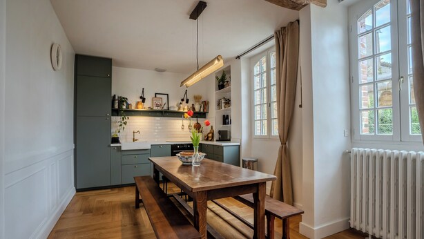 Een keuken met houten vloeren, een houten tafel met banken, een hanglamp en een raam met gordijnen.