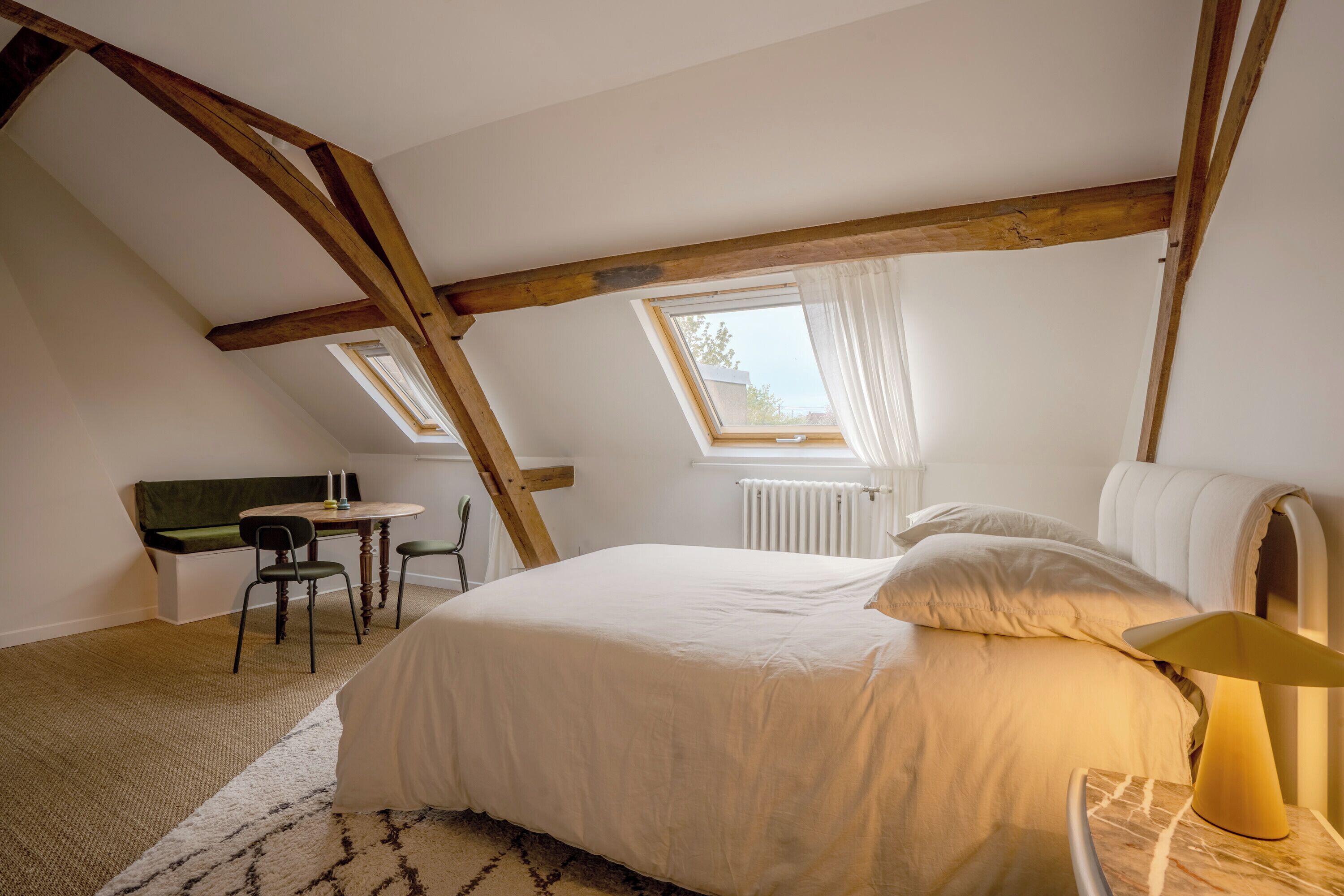 Una camera da letto con soffitto a falde, un letto con biancheria bianca, una lampada da comodino, un piccolo tavolo con sedie e una finestra con tende.
