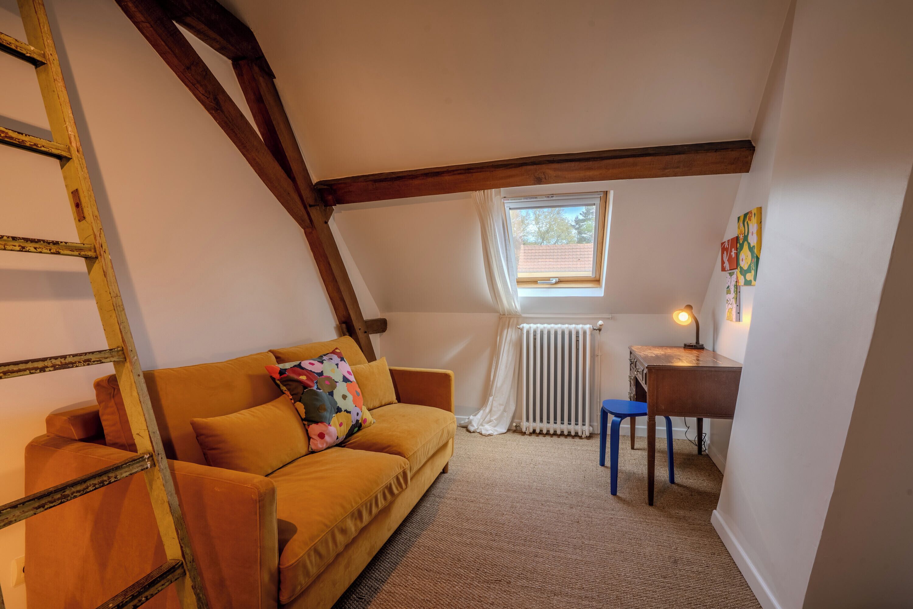 A cozy room with a wooden ladder, a sofa with a colorful pillow, a small table with a lamp, and a window with a view of trees.