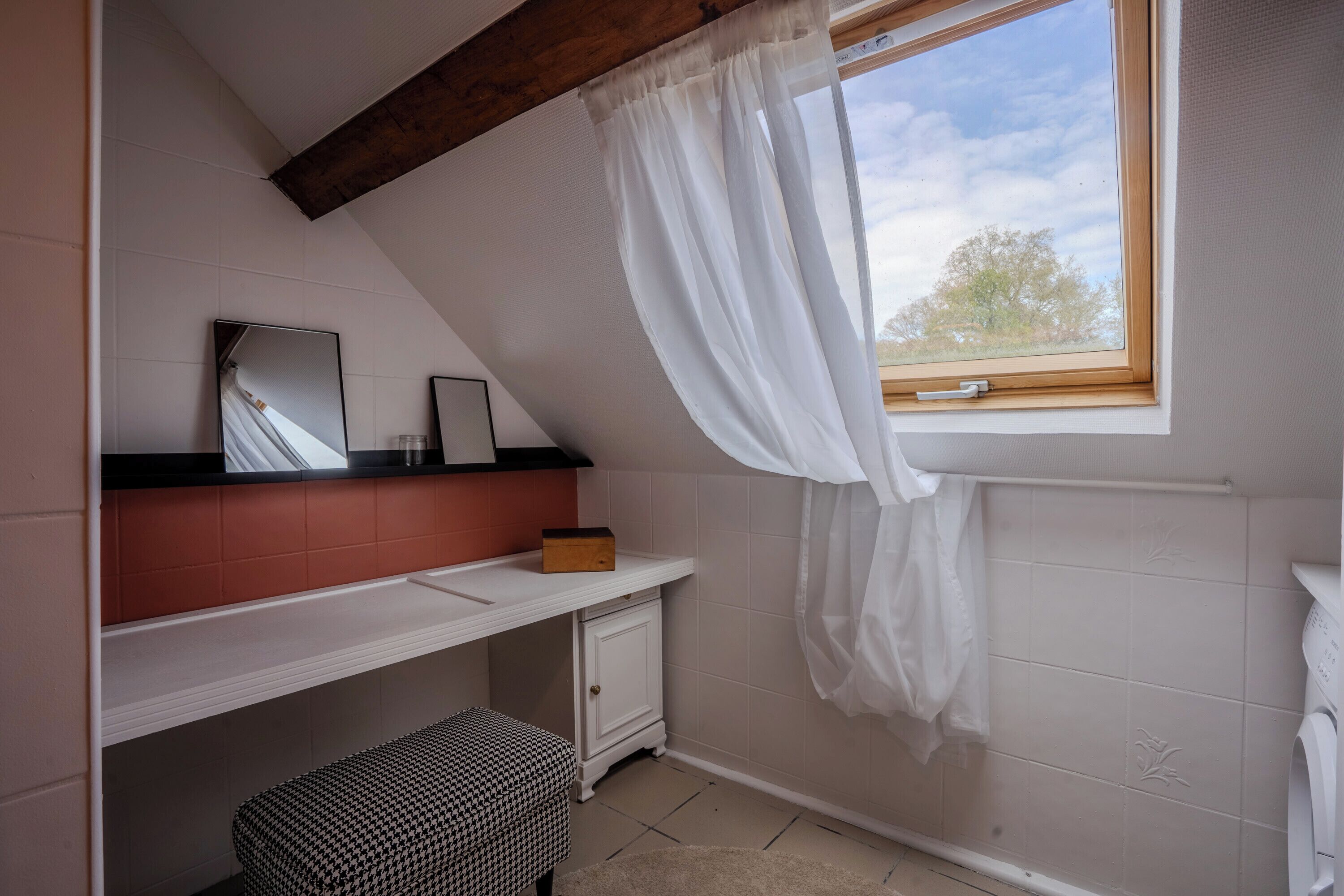 Un baño pequeño y bien iluminado con techo inclinado, una ventana con cortina blanca, un espejo, un lavamanos y un banco.