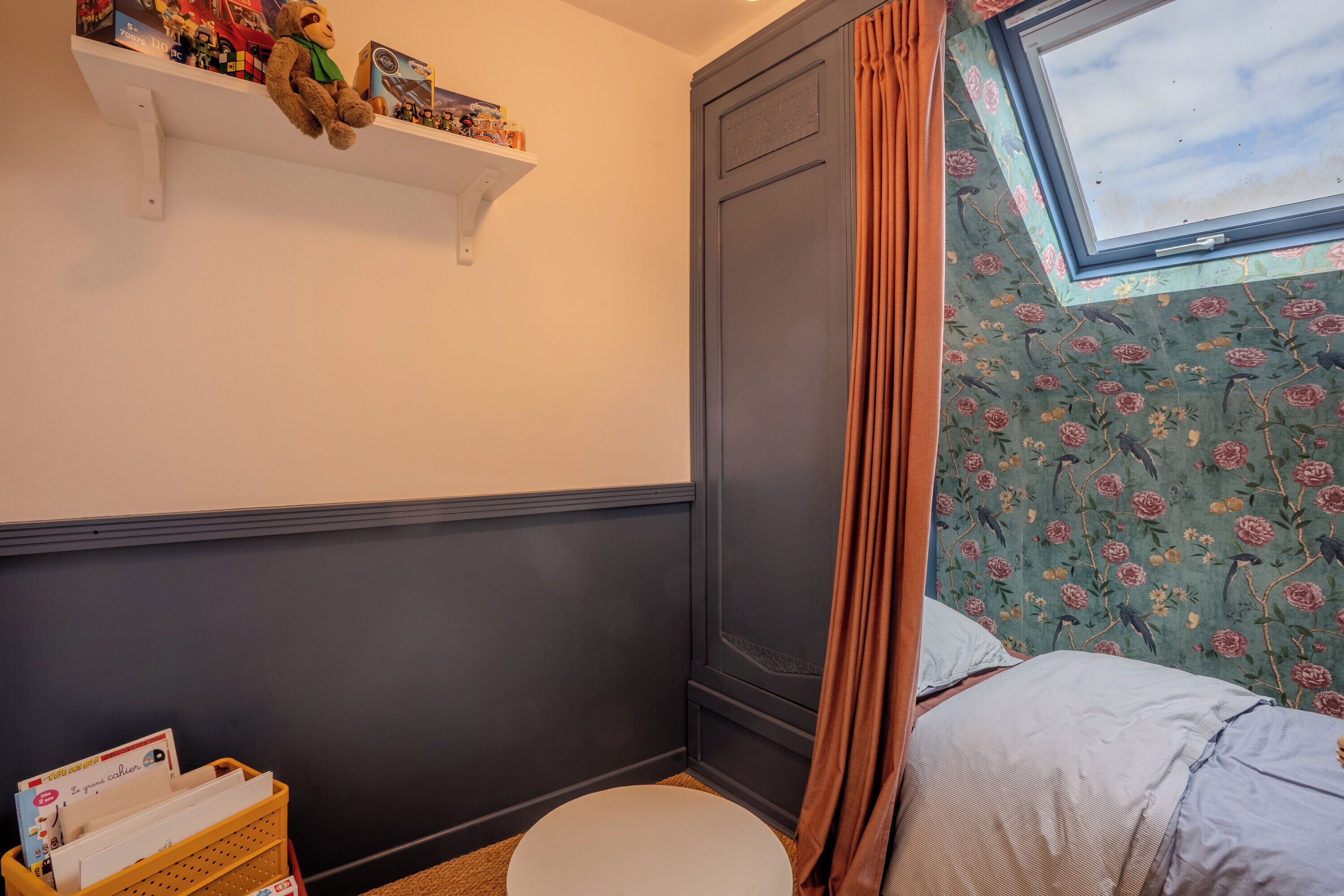 Ein kleiner, gemütlicher Raum mit einem Einzelbett, einer weißen Toilette, einem Regal mit Spielzeug und einem Fenster mit Vorhängen.