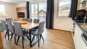 Ein modernes Hotelzimmer mit einem Holztisch, grau gepolsterten Stühlen, einer Küchenzeile und großen Fenstern mit Blick auf eine gebirgige Landschaft.