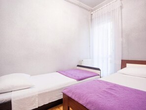 Quarto com duas camas, janela com cortinas leves e radiador.