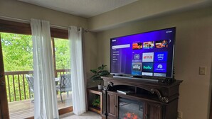 Una sala de estar con una televisión de pantalla plana sobre un mueble de madera, una chimenea, una zona de comedor con una mesa y sillas, y un ventanal corredizo que da a una terraza.