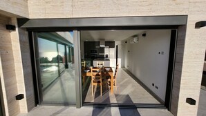 Una cocina moderna con armarios oscuros y una isla central, una zona de comedor con mesa y sillas, y un amplio ventanal corredero que da a una terraza exterior.
