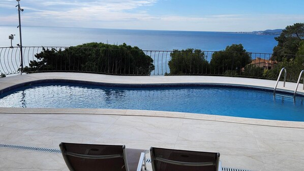 A swimming pool with a view of the sea, surrounded by trees and a metal fence.