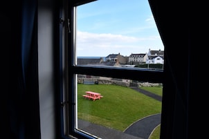 Vista a través de una ventana que muestra un espacio verde con una mesa de picnic roja, un muro de piedra y edificios al fondo.