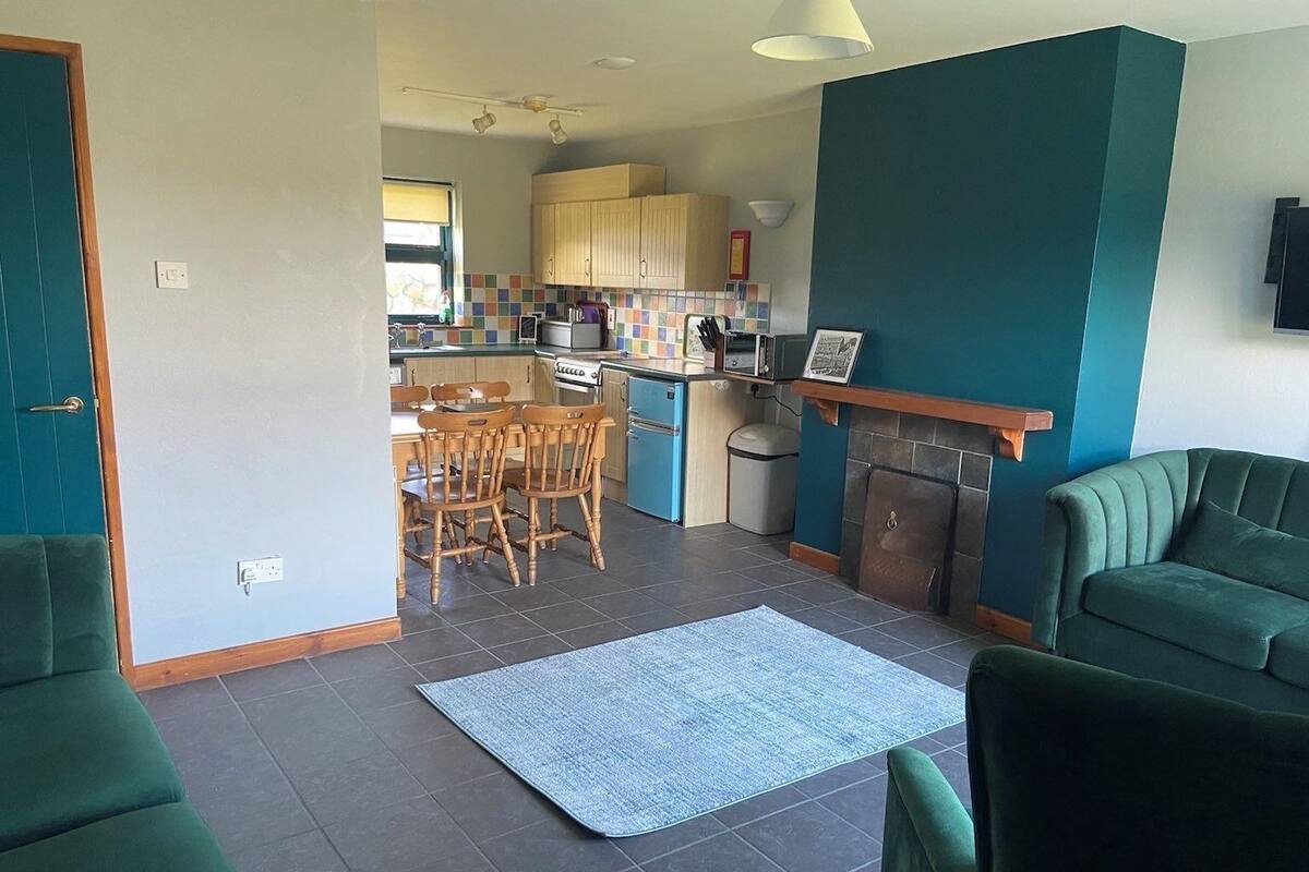 Ein Wohnzimmer mit grünen Sofas, ein Essbereich mit Holzsesseln, eine Küche mit Holzschränken und ein Kamin.
