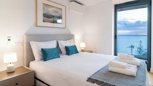 A modern bedroom with a large bed, two pillows, a bedside table with a lamp, a framed picture on the wall, and a view of the sea through a large window. - GuestReady - Acqua Residences - Diamond (Funchal)