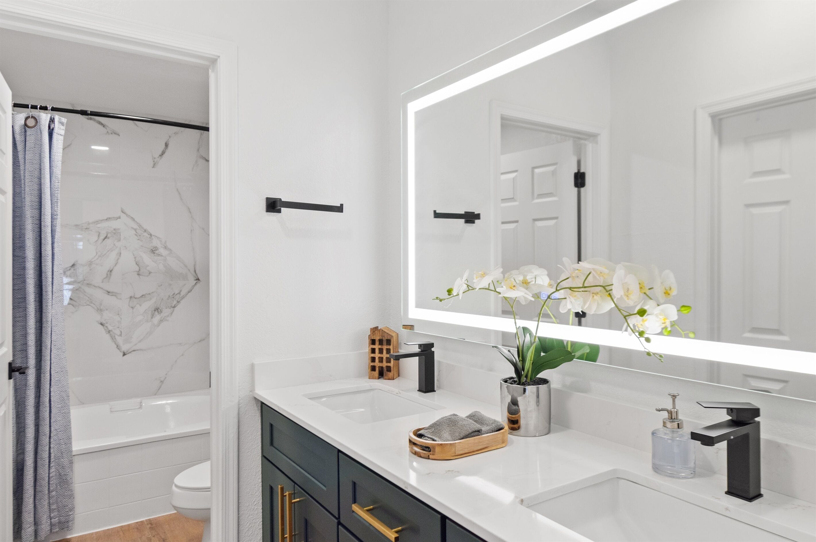 Ein modernes Badezimmer mit einer weißen Marmerdusche, einem großen Spiegel, einem Waschbecken mit Seifenspender und einem Topf mit einer Orchidee.
