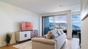 Un salon moderne avec un canapé, un téléviseur à écran plat et une vue sur le paysage urbain grâce à de grandes fenêtres.