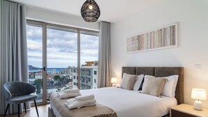 Une chambre d’hôtel avec un grand lit, une chaise, une table de chevet et une vue sur la ville grâce à de grandes fenêtres.