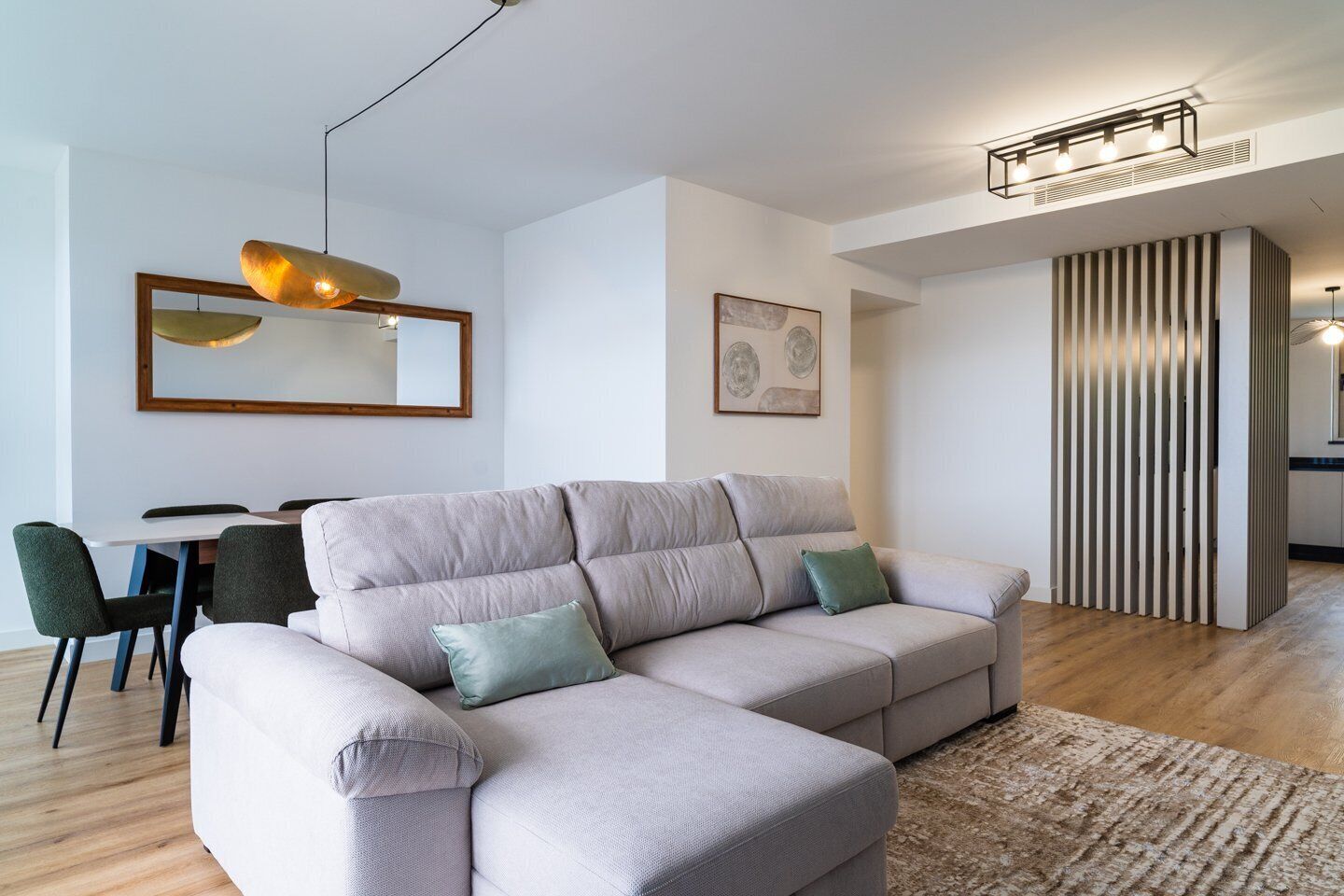 Un salon moderne avec un canapé d’angle gris clair, une table à manger avec des chaises, un luminaire suspendu et une œuvre d’art encadrée au mur.