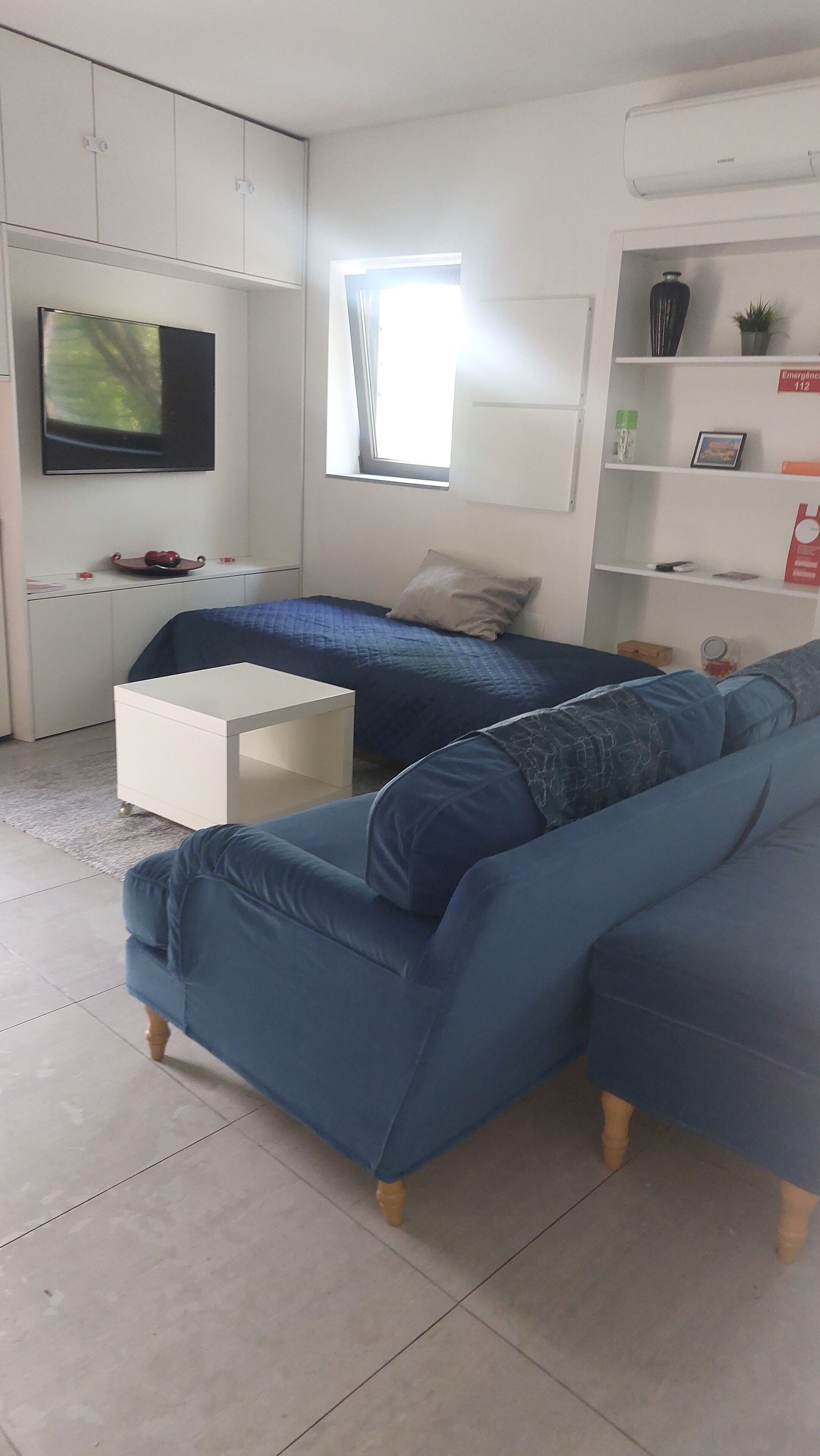 A modern living room with a sectional sofa, a flat-screen TV, a small white table, and a window with natural light.