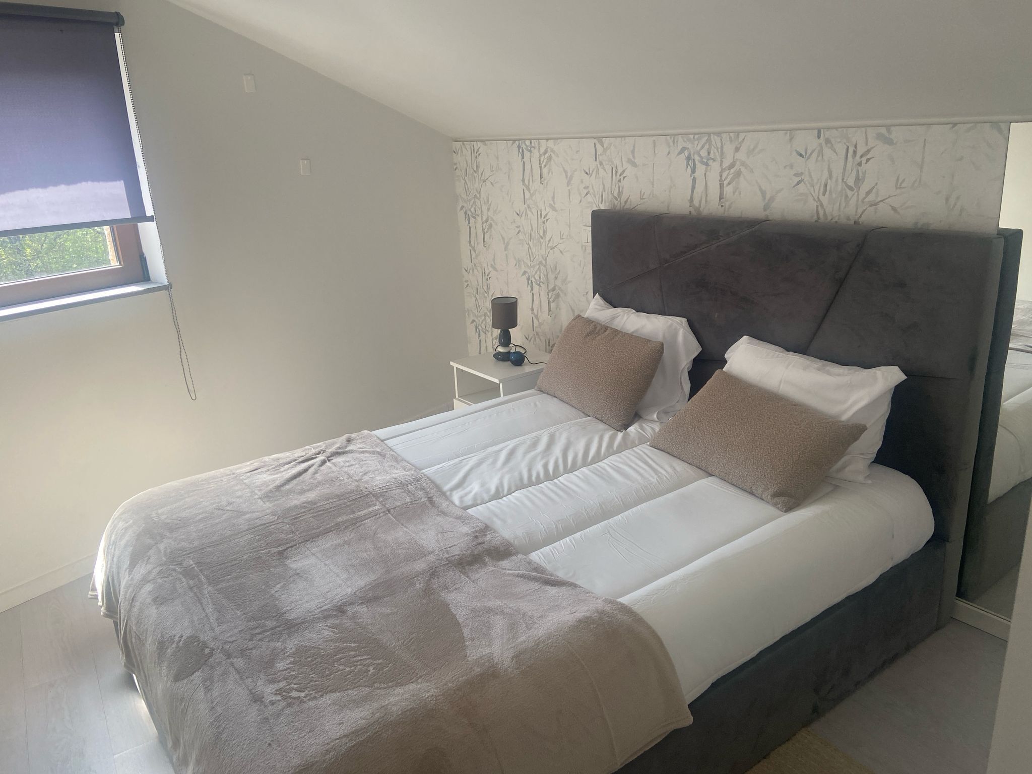A modern bedroom with a large bed, a nightstand, a lamp, and a window with a view.