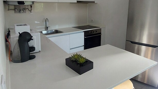 A modern kitchen with white cabinets, a stainless steel refrigerator, a built-in oven, and a small island with stools. - Costa Guest House (Guimarães)
