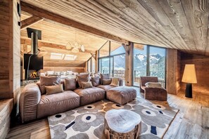 A cozy log cabin interior with a large sectional sofa, a fireplace, and a view of mountains through large windows. - Jolimont - Elegant Alpine Chalet in Châtel (Châtel)