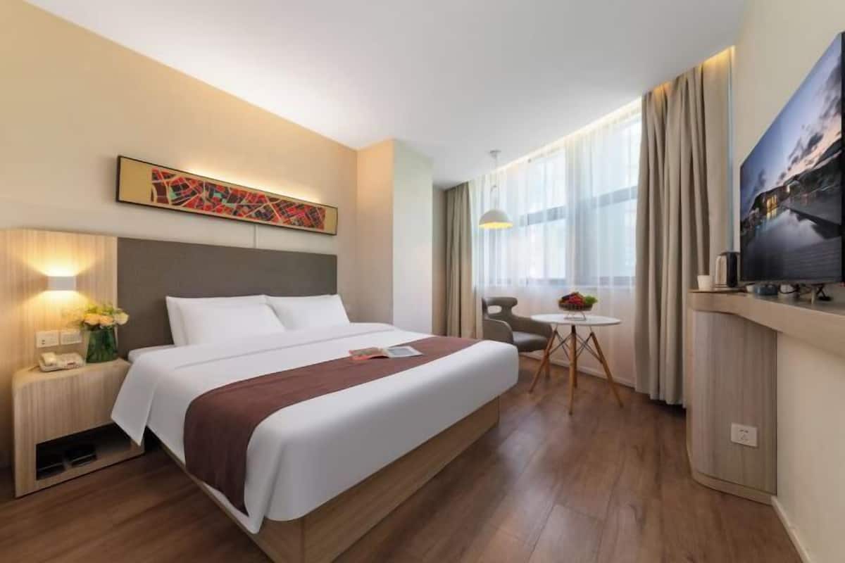 A hotel room with a large bed, a small table, a chair, a TV, and a painting on the wall.