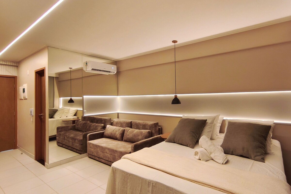 A modern hotel room with a large bed, a sofa, a mirror, and a ceiling-mounted air conditioner.