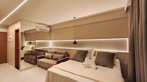 A modern hotel room with a large bed, a sofa, a mirror, and a ceiling-mounted air conditioner. - Malia Beach Experience  (Ipojuca)