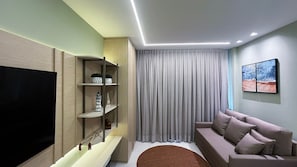 A modern living room with a flat-screen TV, a sofa, a coffee table, a TV stand with shelves, and a large window with curtains. - Malia Beach Experience  (Ipojuca)