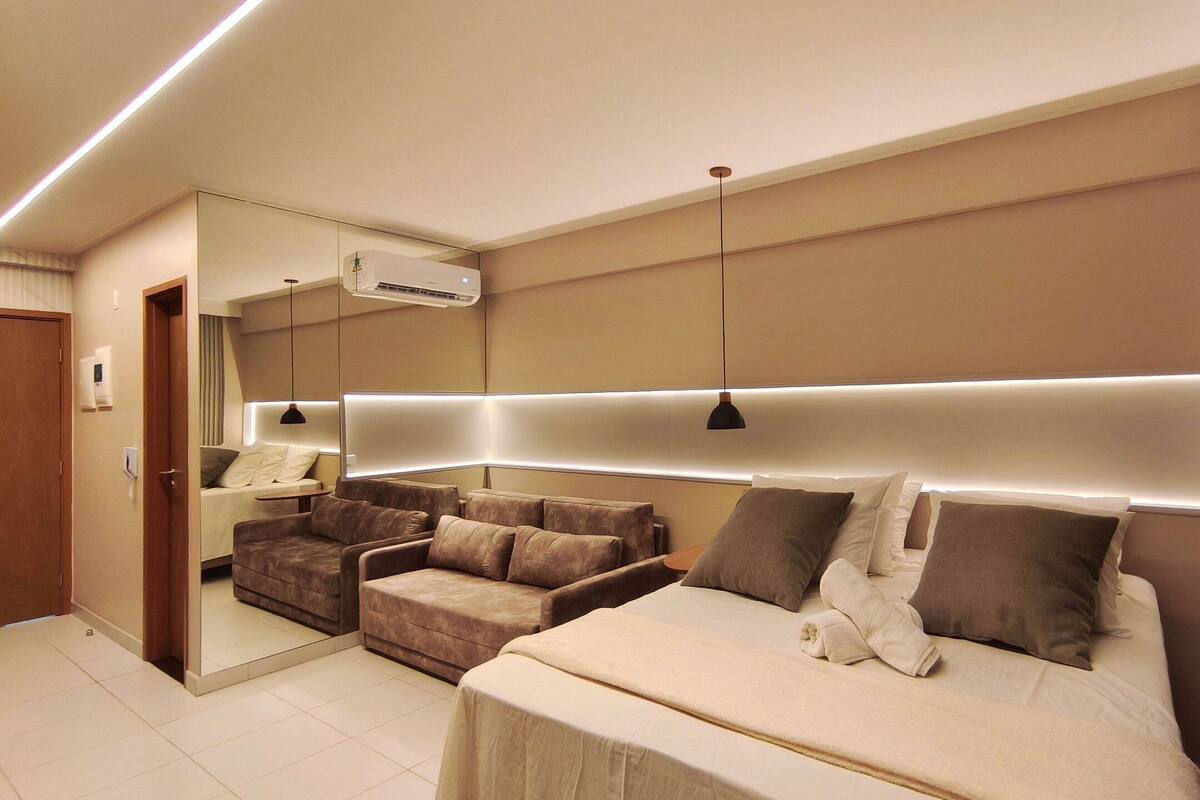 A modern hotel room with a large bed, a sofa, a mirror, and a ceiling-mounted air conditioner.