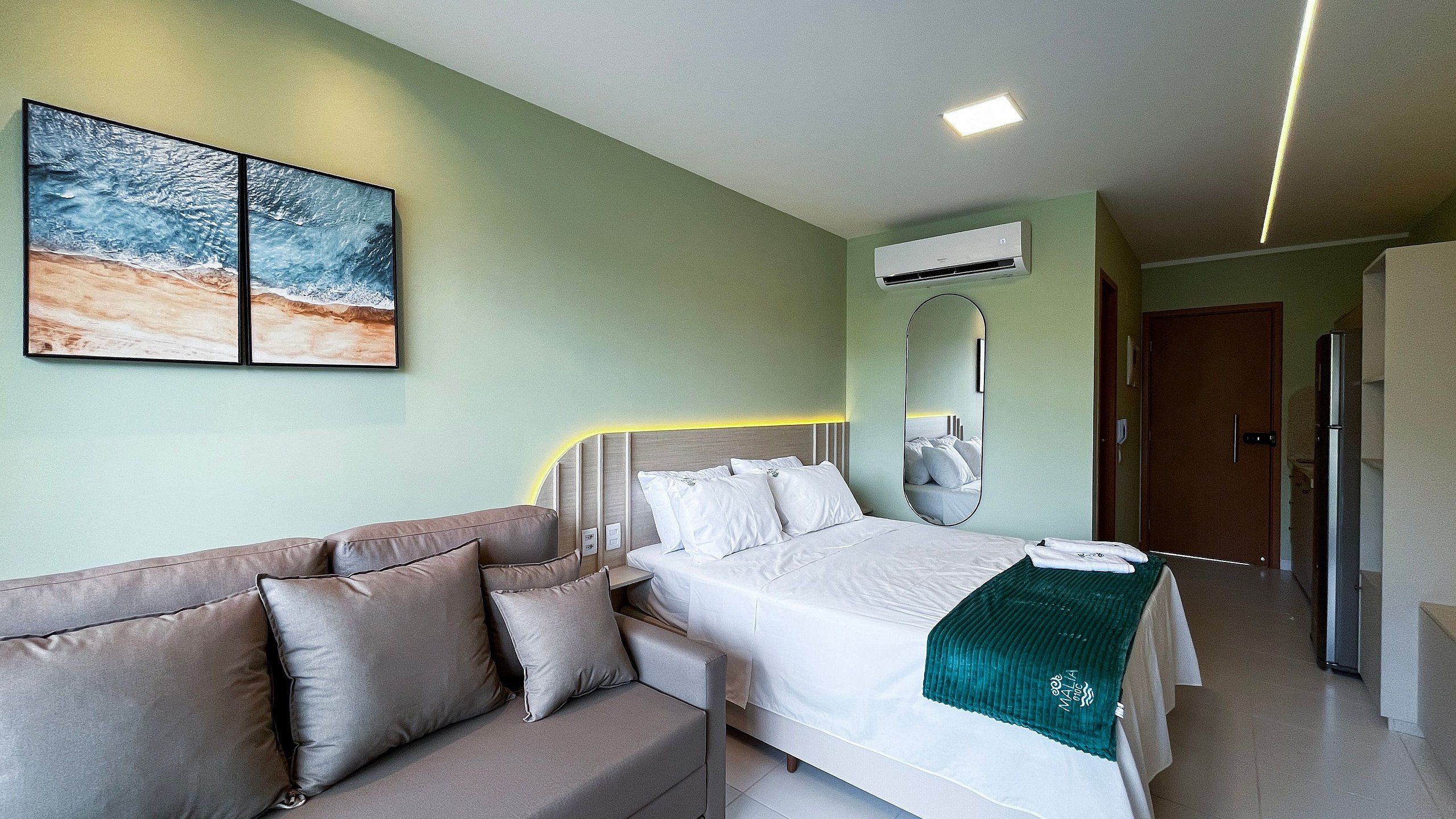 A hotel room with a bed, a sofa, a wall-mounted mirror, an air conditioning unit, and a door.