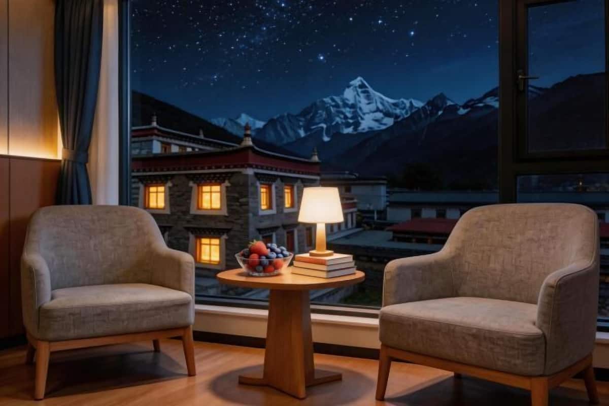 A room with two armchairs, a round wooden table with a lamp, and a view of snow-capped mountains through a window.