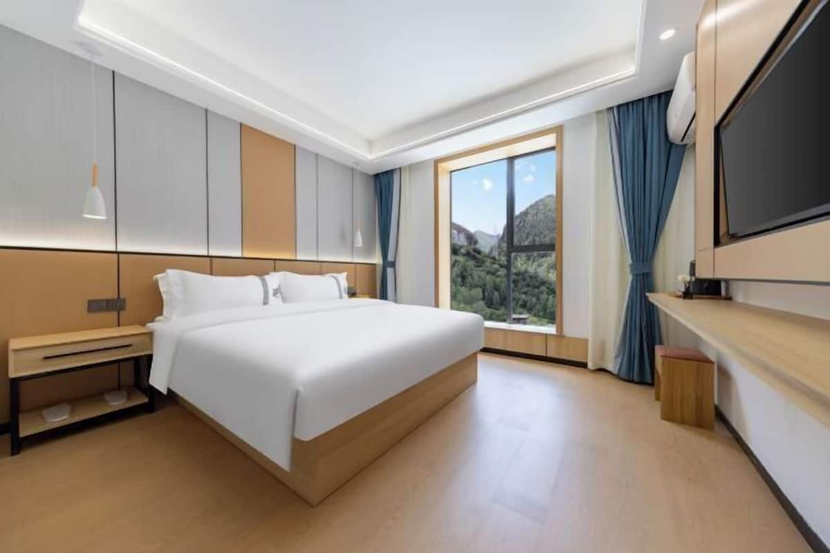 A modern hotel room with a large bed, a TV, a window with a mountain view, and a wooden floor.