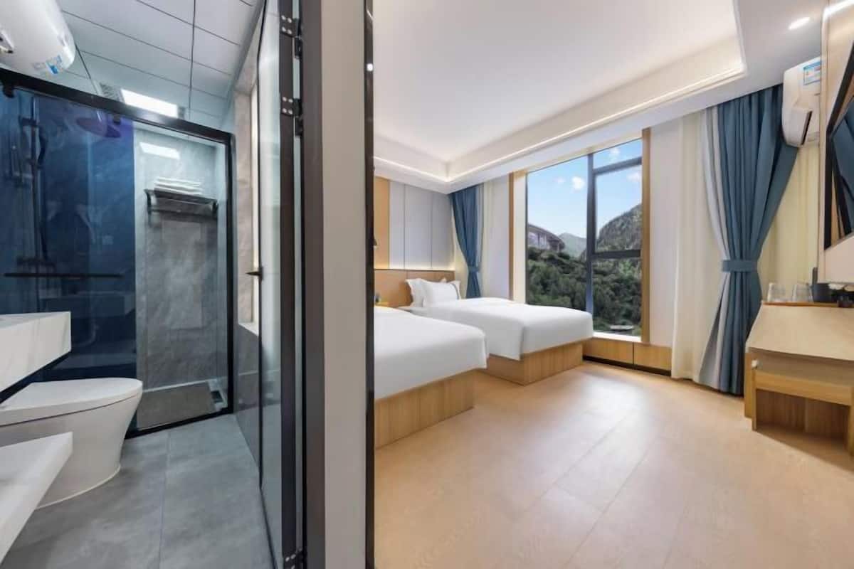A modern hotel room with a glass shower enclosure, two beds, a large window with a view, and a wooden bench.