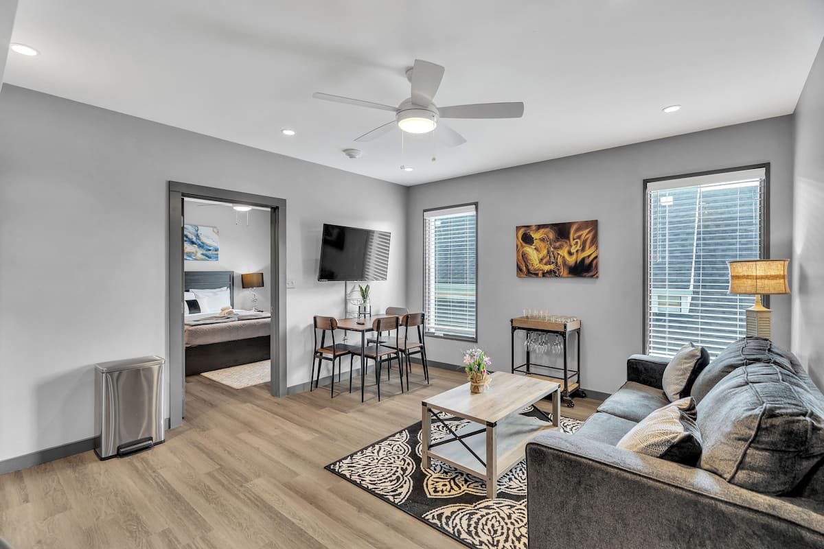 A modern living room with a ceiling fan, a dining area with a table and chairs, and a large window with blinds.