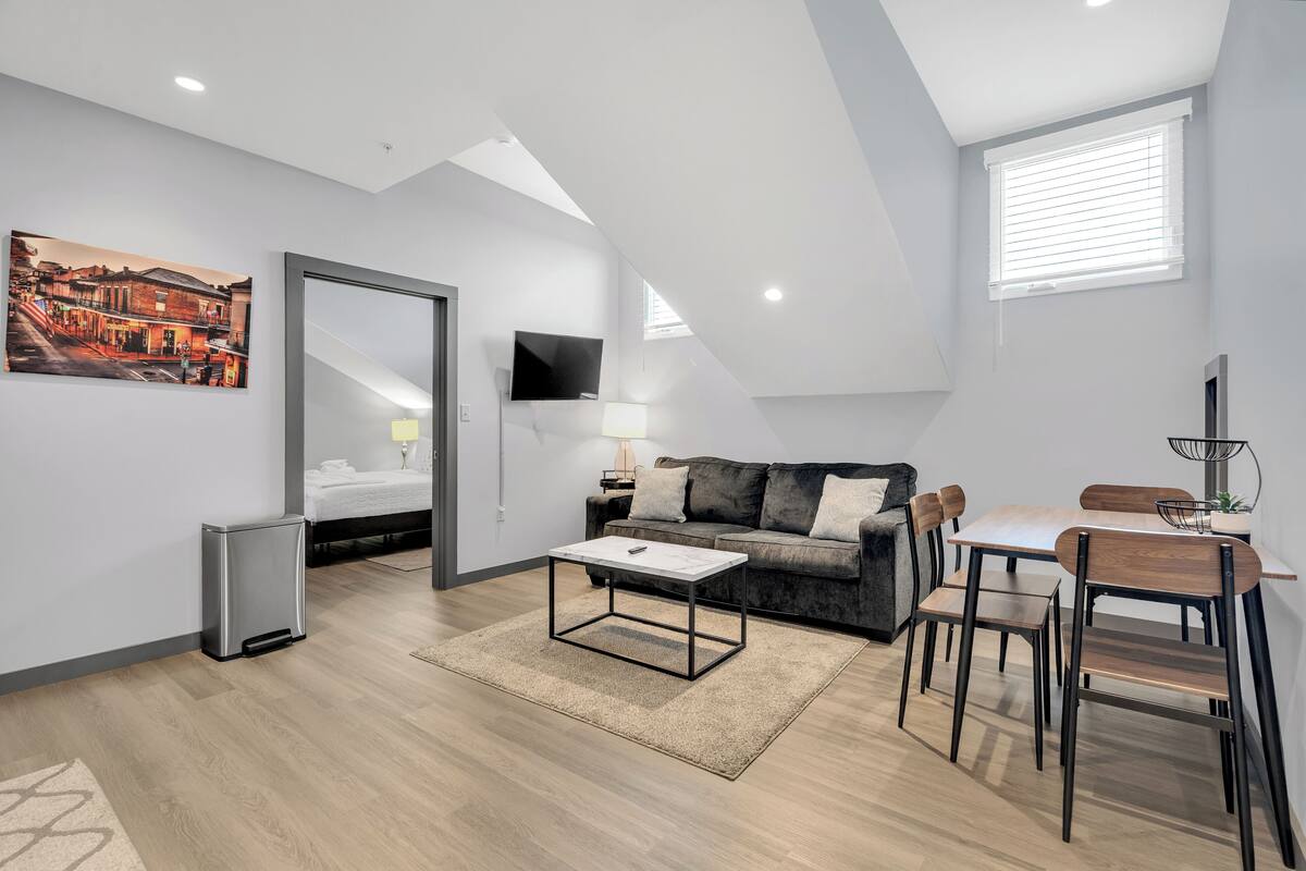 A modern loft apartment with a dining area, a sofa set, a TV mounted on the wall, and a painting of a street scene.
