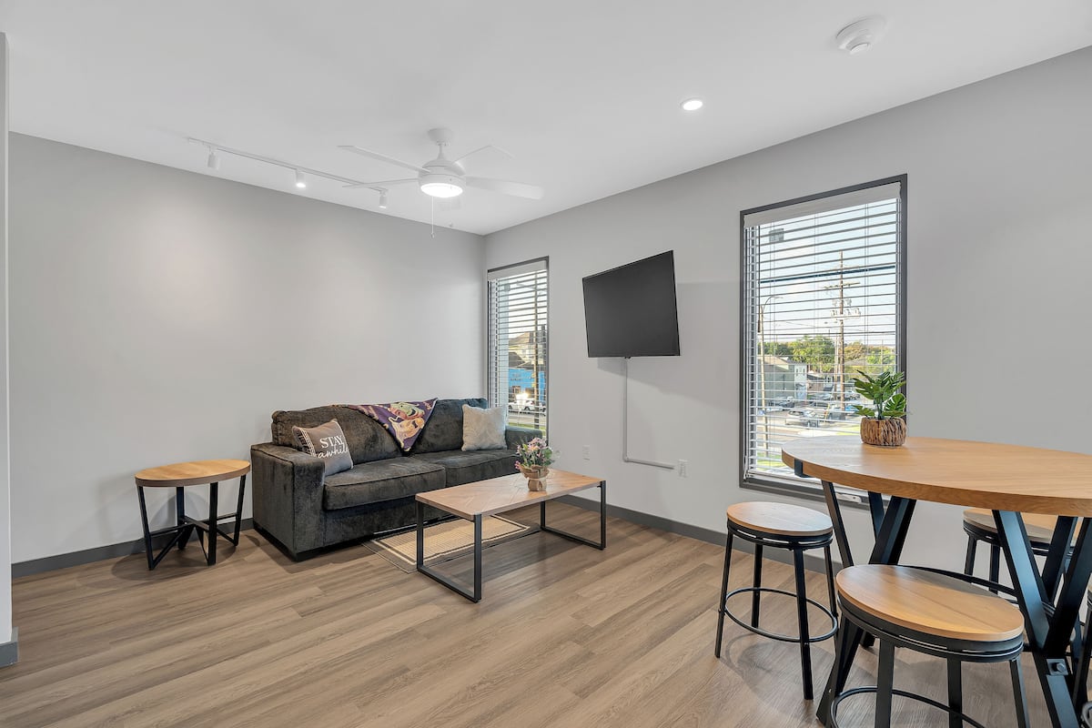 A modern living room with a grey sofa, a wooden coffee table, a TV mounted on the wall, and large windows with blinds.