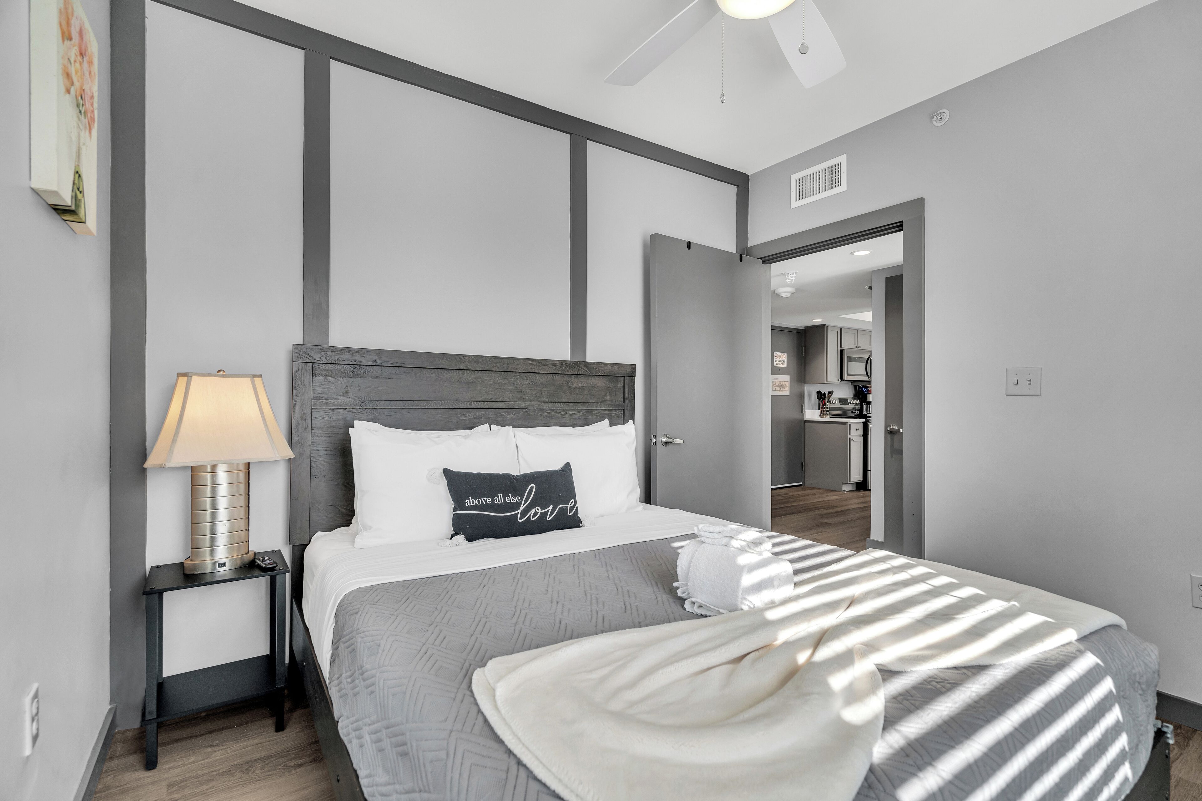 A modern bedroom with a bed, a nightstand, a lamp, and a view into the kitchen.