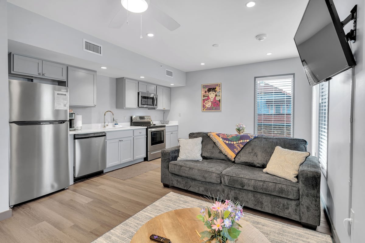A modern kitchen with stainless steel appliances, a grey sofa, and a flat-screen TV mounted on the wall.