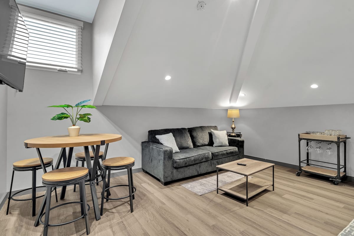 A modern loft apartment with a high ceiling, a grey sofa, a wooden dining table with stools, and a small side table with a lamp.