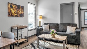 A modern living room with a grey sofa, a coffee table, a dining table, and a wall-mounted art piece. - New Orleans Bywater Apartments (New Orleans)