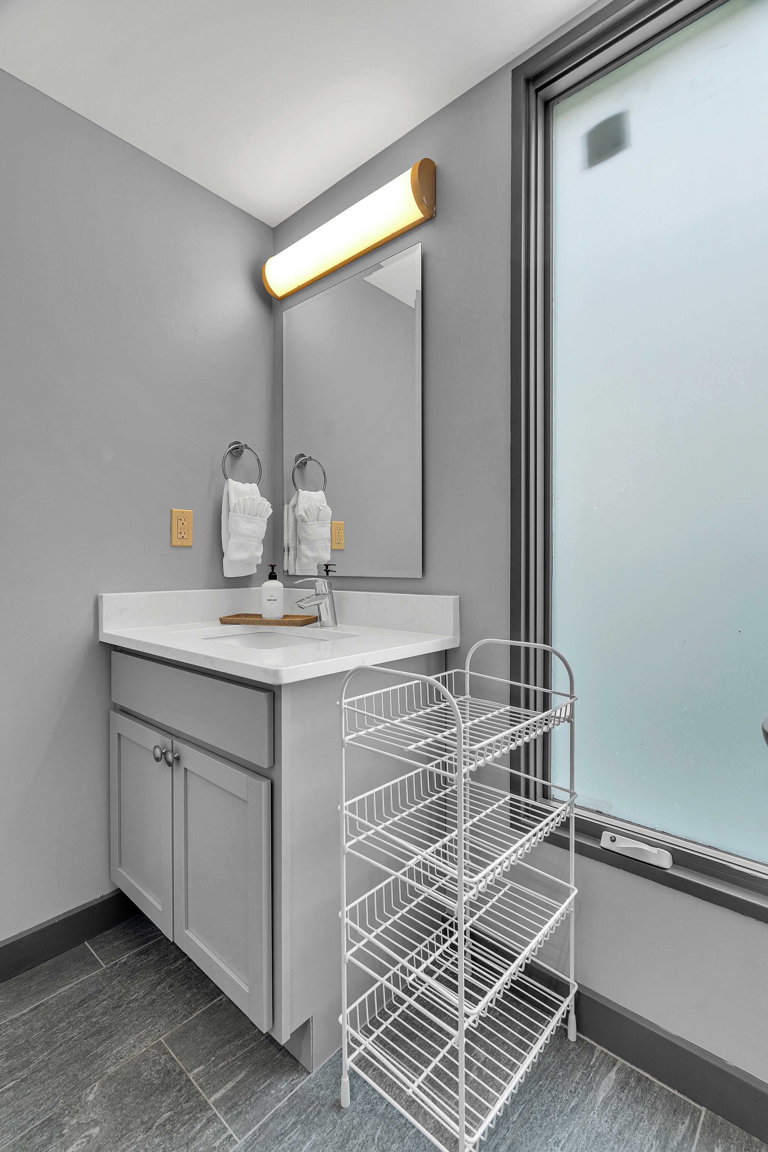 Une salle de bain moderne avec un mur gris, un meuble-lavabo blanc, un miroir avec éclairage intégré et une étagère en métal.
